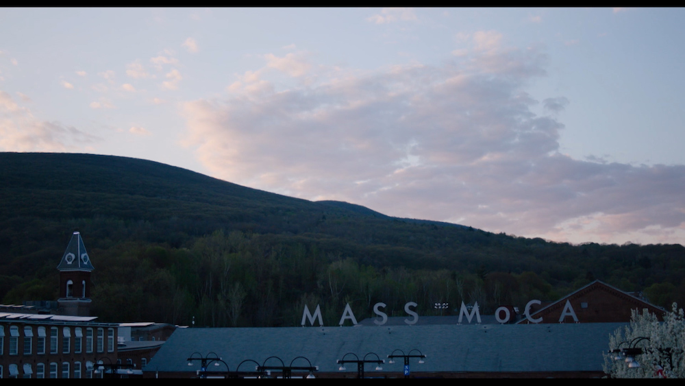 Film still shows the buildings and signage of MASS MoCA against mountains and sky at dusk or dawn.