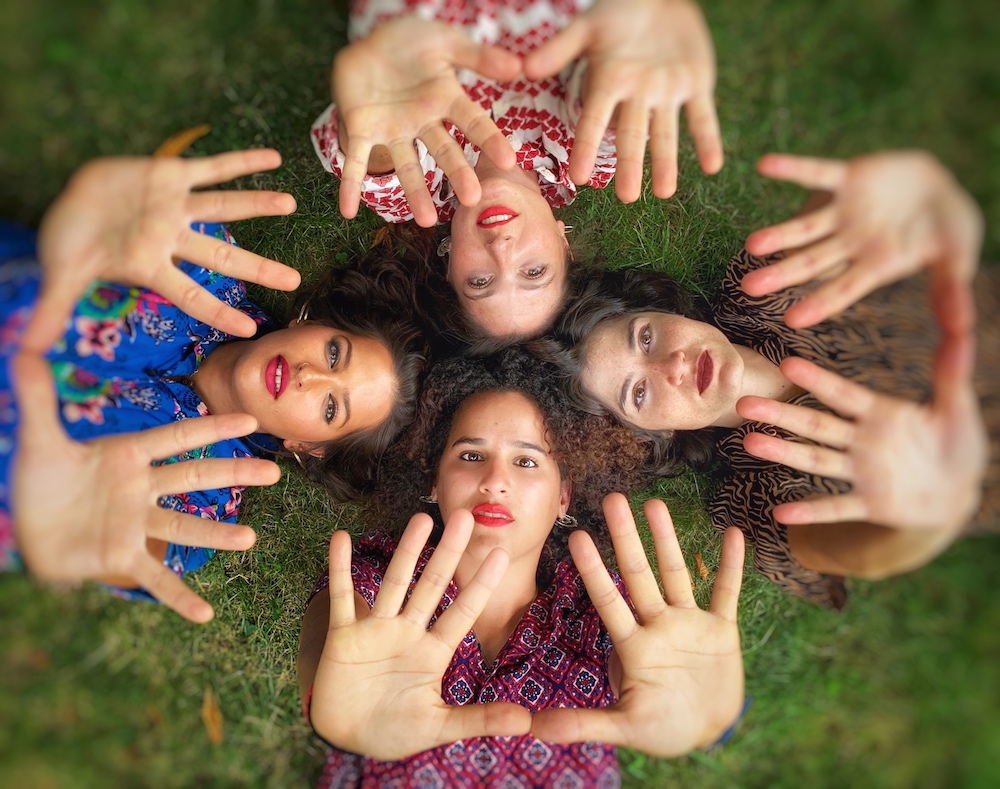 Seen from above, four young women lay in grass with their heads together and their hands extended toward the camera.