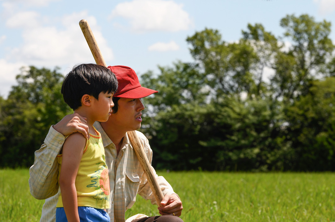 Minari film still, with Steven Yuen's character kneeling and holding a shovel and the other hand on the shoulder of on-screen son. The background shows grass and trees. 