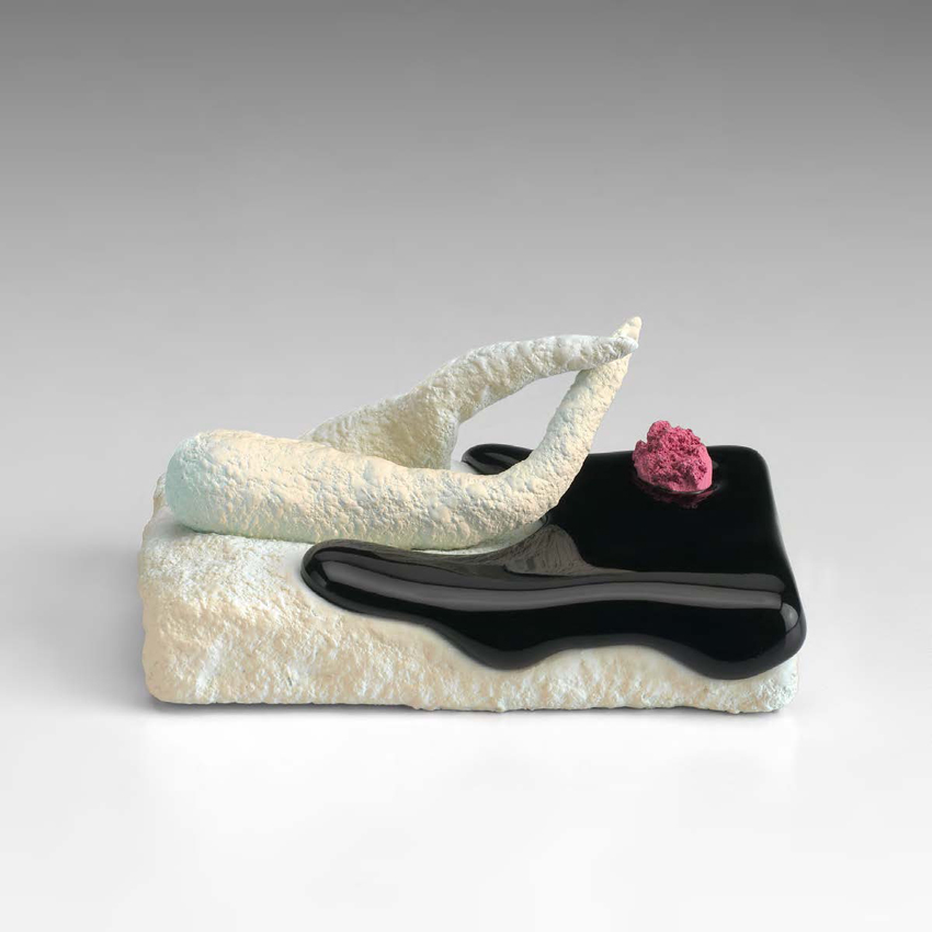 A small ceramic sculpture comprises a rectangular slab with two protruding, crossed, leg-like forms, a shiny black puddle that oozes down the sides, and a red flower-like tuft.