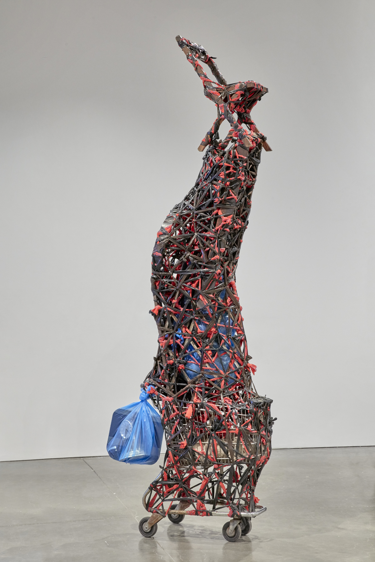 A sculpture made of various objects piled on top of a metal shopping cart, tightly wrapped and bound with twisted red and black plastic, culminating in a simple folding chair at the very top.