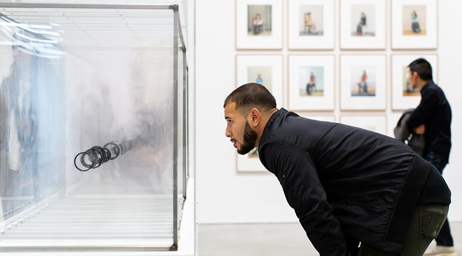 A man in a black jacket leans forward to closely observe a transparent display with circular objects inside. A gallery wall with framed photos is in the background. Another person stands nearby, also viewing the art.