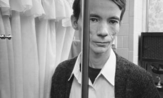 A black-and-white photograph of a light-skinned man reflected in two mirrors with pale tiles and a shower curtain behind him.