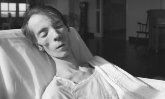 A black-and-white photograph of a pale emaciated man asleep in a hospital bed with bright sunlight shining on his face.