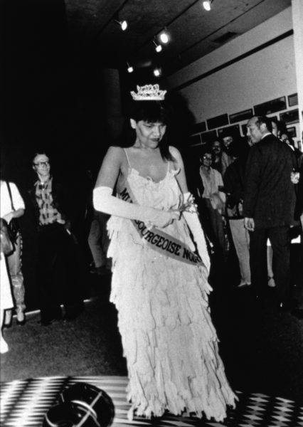 A black-and-white photograph of a Black woman in a beauty pageant gown, crown, gloves, bouquet, and sash walking across a crowded gallery while looking down at the floor.