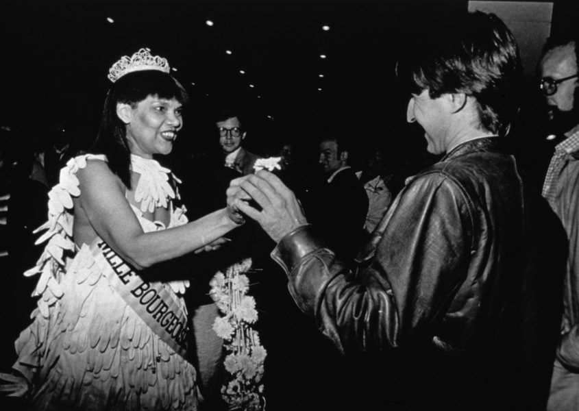A black-and-white photograph of a Black woman in a beauty pageant gown, crown, gloves, bouquet, and sash extending appearing to hand a small flower to a light-skinned man in a leather jacket.