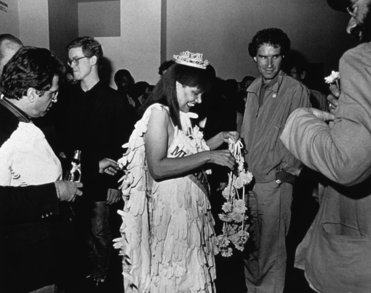 A black-and-white photograph of a Black woman in a beauty pageant gown, crown, gloves, bouquet, and sash standing in the center of a crowd,adjusting her bouquet while smiling and, looking down.