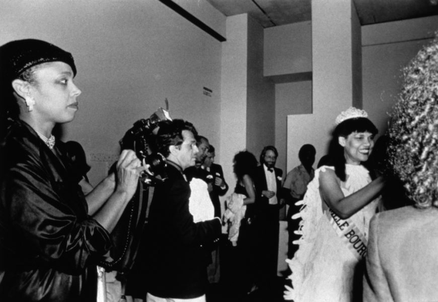 A black-and-white photograph shows a Black woman in a beauty pageant gown, crown, gloves, bouquet, and sash in the bottom right corner of the image greeting a stranger as others wait to greet and photograph her. 