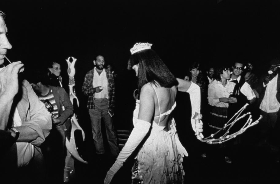 A black-and-white photograph of shows a Black woman in a beauty pageant gown, crown, and gloves whipping a white cat o’ nine tails behind her in a crowded gallery.