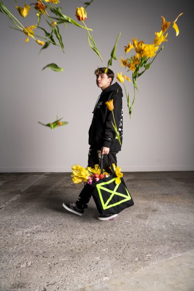 A young man dressed all in black walks looks toward the camera mid-stride as yellow tulips fly toward him.