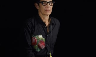 A color photograph shows the choreographer Elizabeth Streb, a pale, middle-aged woman with spiky dark hair, dark-rimmed glasses, black clothing with a large embroidered floral design on the chest, and a serious expression, seated in a three-quarter profile against a black background.