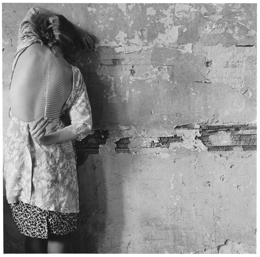A black-and-white photograph of a woman leaning her head against a wall, holding a fish spine in front of her own exposed back.