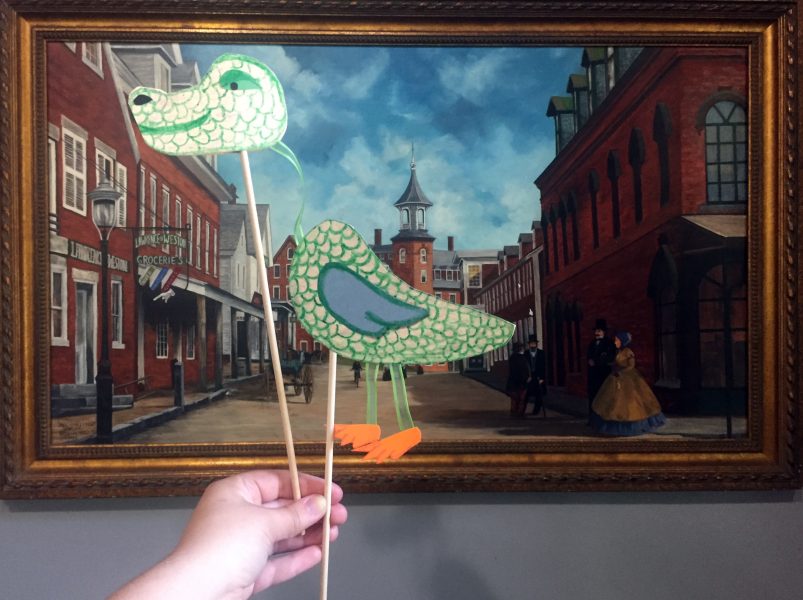 Paper puppet held in front of a painting of a street