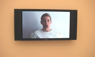 A black video monitor with attached speakers on either side shows a still of a pale-skinned woman in a white t-shirt in mid-speech as she faces the viewer.