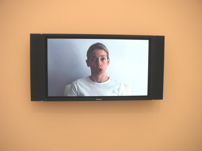 A black video monitor with attached speakers on either side shows a still of a pale-skinned woman in a white t-shirt in mid-speech as she faces the viewer.