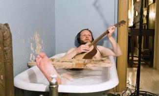 A video still shows the artist, a white man, in a bathtub, wearing headphones, and plays the guitar, and apparently nude.