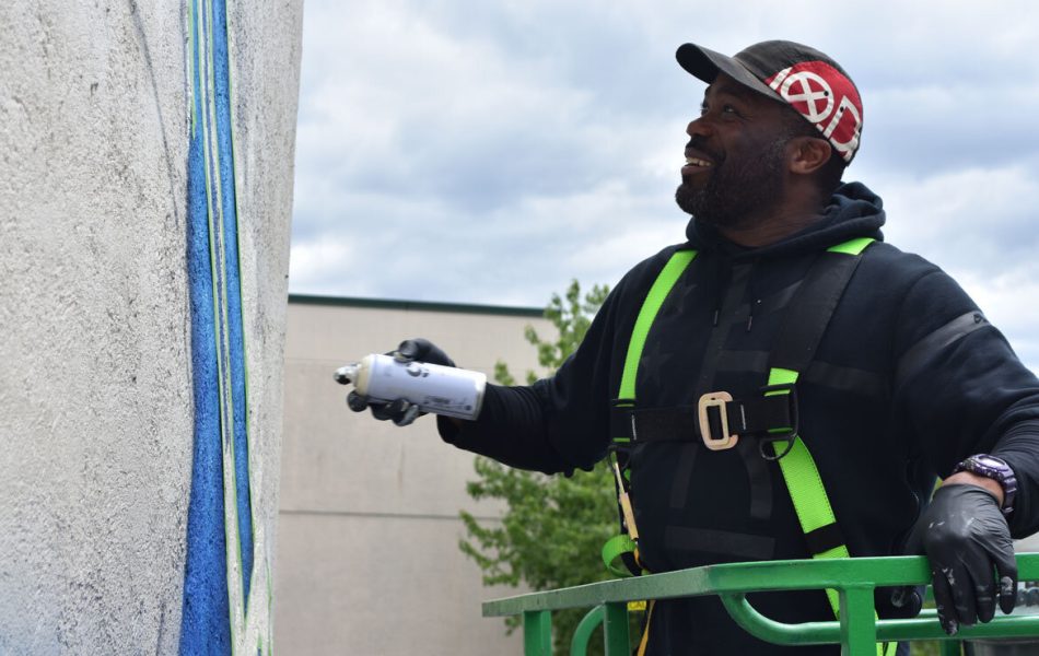 A man wearing a harness stands in a lift and spraypaints a wall