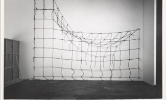 A black-and-white photograph of an installation of brown rope knotted in a grid and hung on a white gallery wall, with the top right corner detached and hanging down.