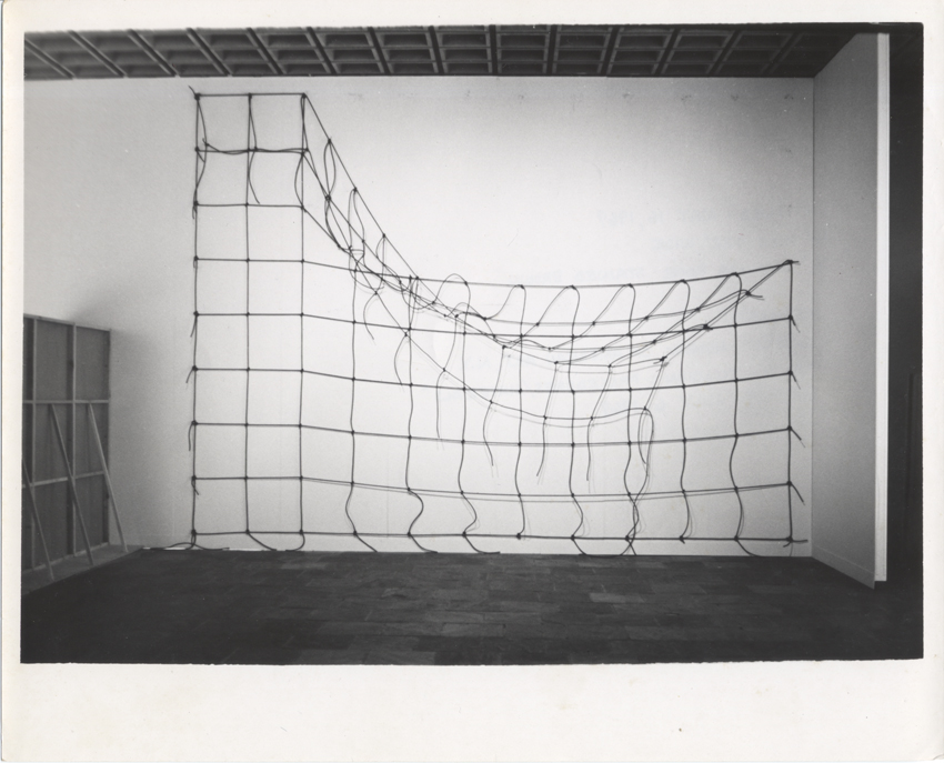 A black-and-white photograph of an installation of brown rope knotted in a grid and hung on a white gallery wall, with the top right corner detached and hanging down.