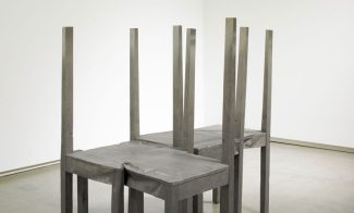 A stainless-steel sculpture of four backless, chairs connected together in the shape of an "L" in an empty gallery.