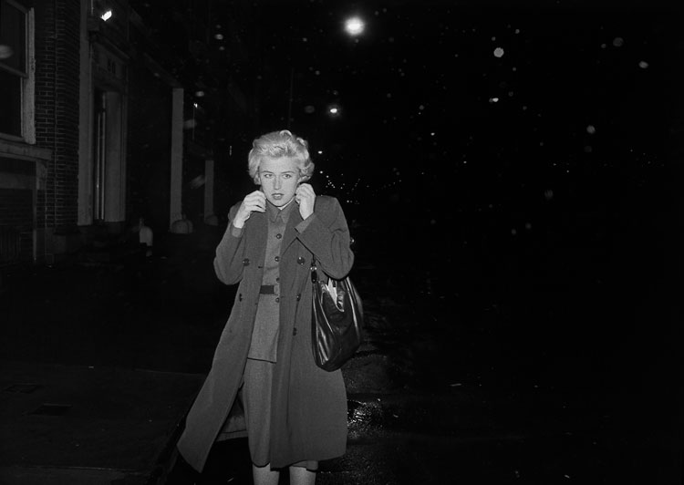 A black-and-white photograph of the artist on a very dark urban street, with blond hair and a dress skirt and blouse under a trench coat that she pulls closed around her neck.