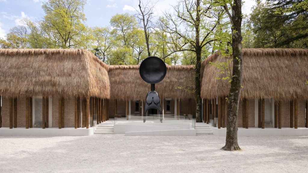 A thatch-roofed building with brick walls and large wooden pillars surrounds a sculpture resembling a figure with a disc-shaped head, set among trees and a gravel pathway under a partly cloudy sky.