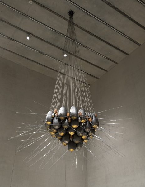 A large, suspended sculpture made of black and gold forms clustered together, with numerous thin metal rods extending outward in all directions, resembling an abstract starburst. It hangs from the ceiling in a minimalist gallery setting.