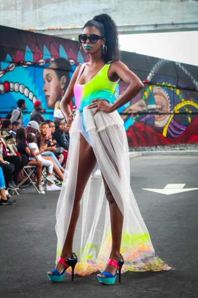 A model in a neon-colored swimsuit and chiffon train posing in an outdoor runway show