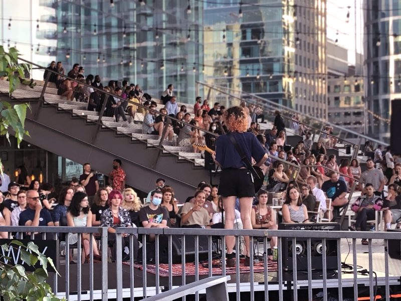 An audience sitting outside the ICA and on the grandstand watch a musical performer on a stage with their back to the camera