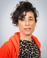 A headshot of choreographer Netta Yerushalmy. She has dark curly hair pulled back and an open orange hoodie over a leopard-print top.