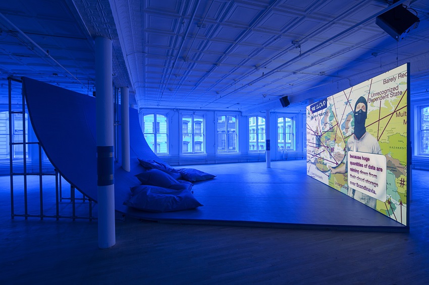 An installation photo cast in blue light of a large video screen showing a masked man in front of a map of Europe, opposite a curved wall-to-ceiling quarter pipe with lounge seating.