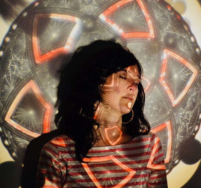 A light skinned woman with dark hair and a projection of a mandala design over her face.