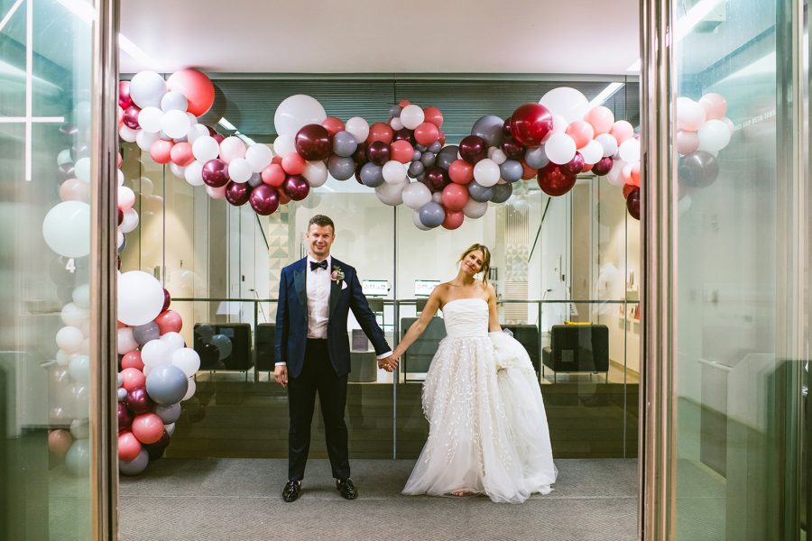 A bride and groom hold hands in the ICA's glass elevator, full of a garland of colored balloons.