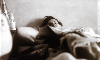 A tinted photograph of the artist Mark Morrisroe, a light-skinned man with dark hair, lying in bed, taken from near the foot of the bed, with just his face peering over thick blankets that obscure his body.