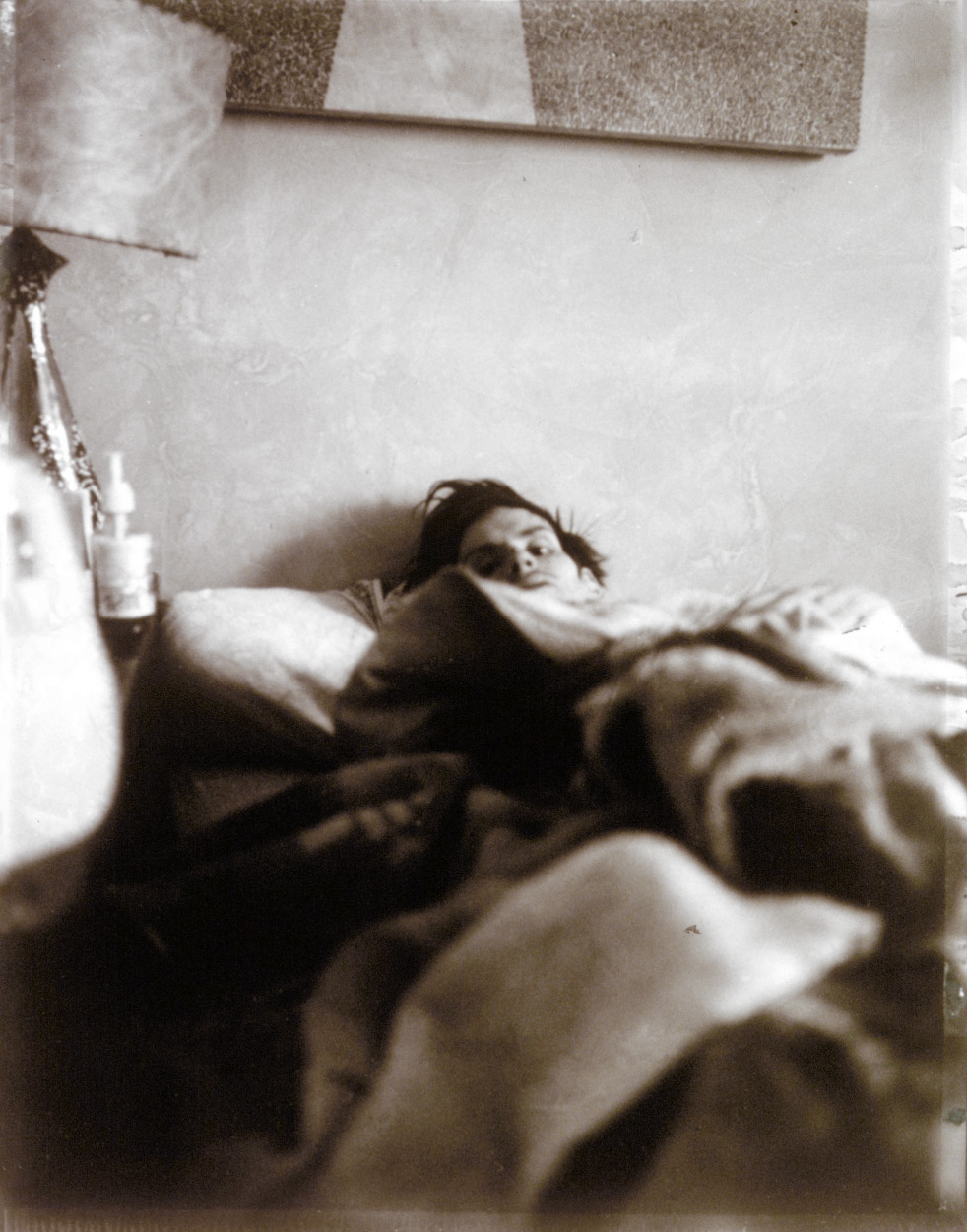 A tinted photograph of the artist Mark Morrisroe, a light-skinned man with dark hair, lying in bed, taken from near the foot of the bed, with just his face peering over thick blankets that obscure his body.