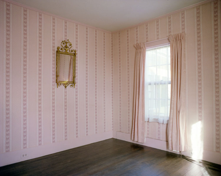 A color photograph of the corner of an empty room with a gold mirror, pink curtains, and decorative pink wallpaper.