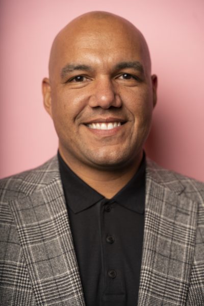 A man in a brown tweed jacket in front of a pink wall.