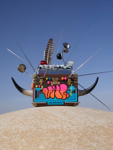 A repurposed guitar amp sits atop a dry, desert-like surface, and is adorned with spikes, animal horns, feather, and other metallic objects with a colorful spray paint on the front surface.