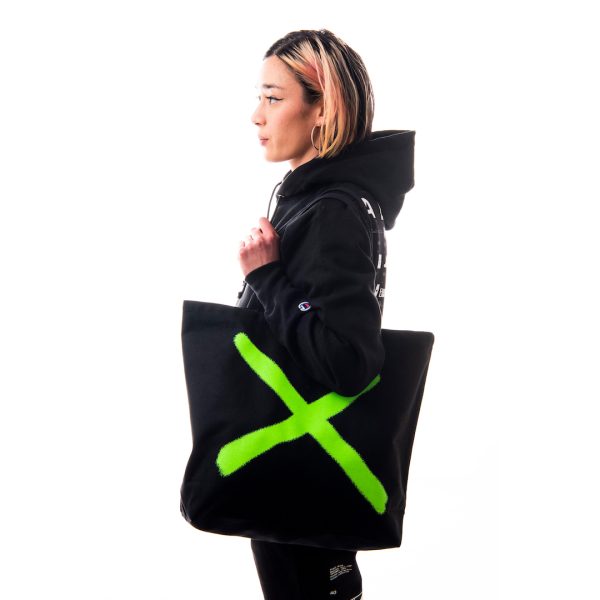 Profile view of light-medium skinned model holding a black tote bag with a large neon green X. 