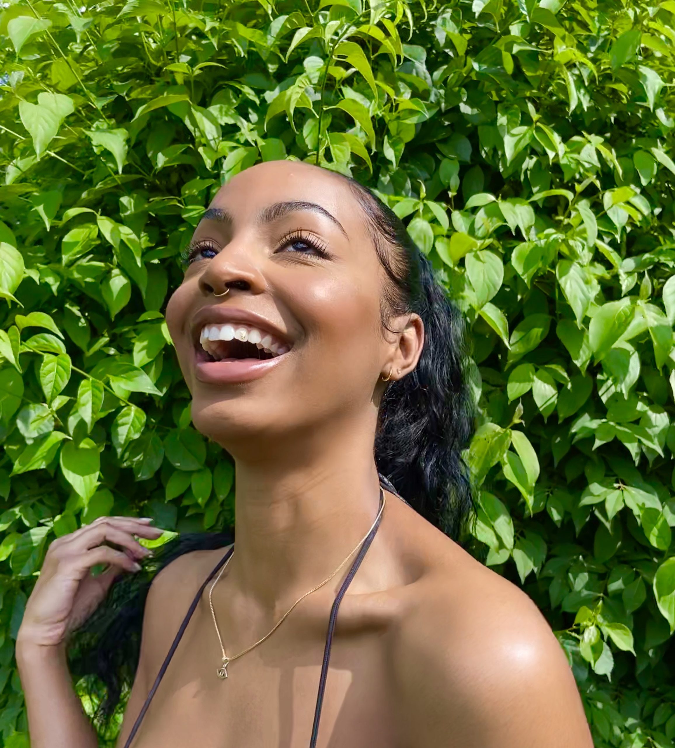 A medium dark-skinned women smiling and looking up with greenery in the background.