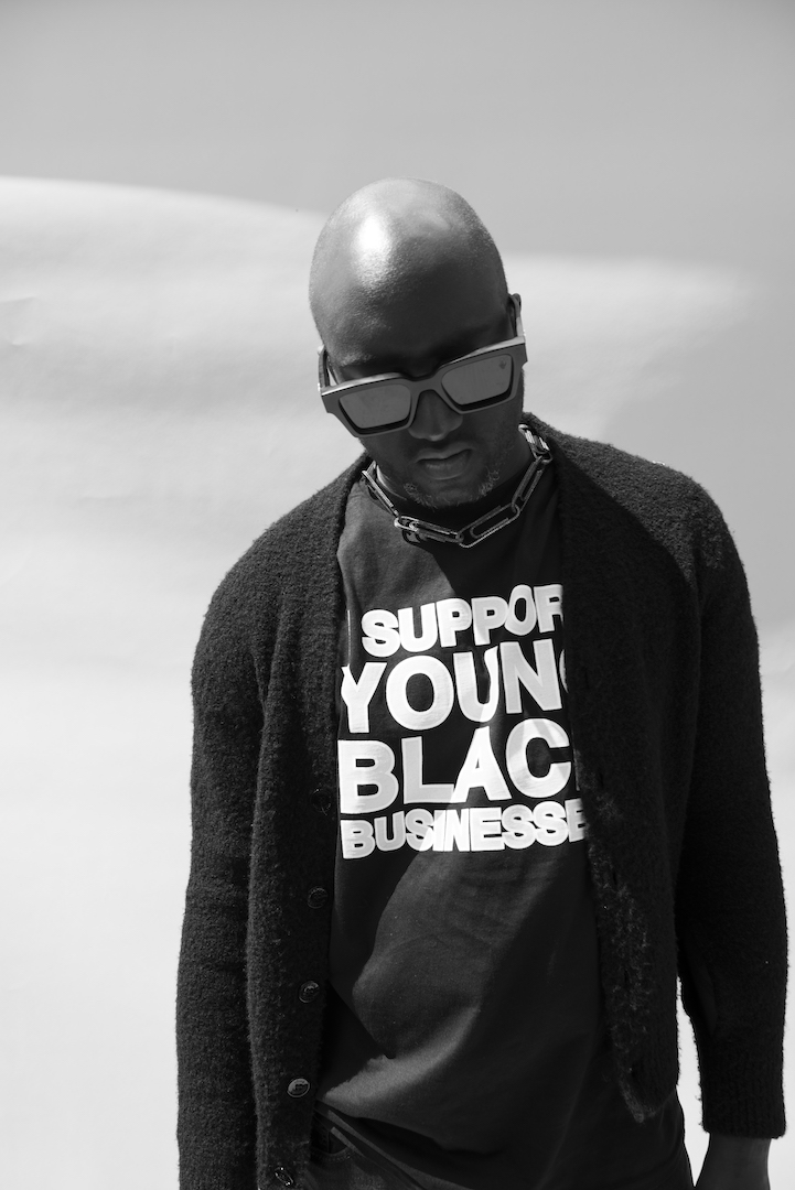 Black-and-white portrait of Virgil Abloh, an African-American man looking down wearing sunglasses with a cardigan over a t-shirt that reads