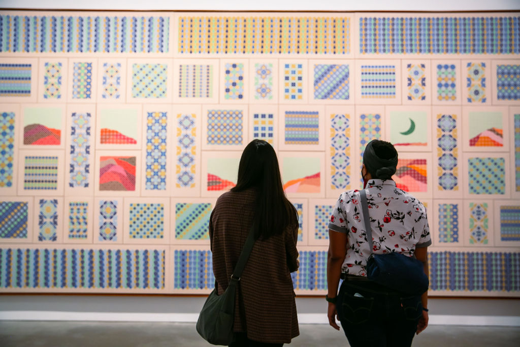 Two visitors look at a large rectangular tapestry with many smaller panels of geometric patterns and landscape imagery
