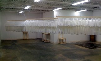 Dozens of white T-shirts hang from hangers attached to three wooden beams suspended from the gallery ceiling.