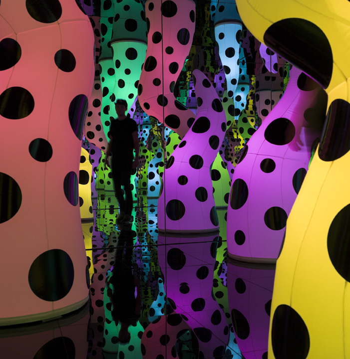 An installation of a polka-dotted neon colored tentacle-shaped arms that extend from the ceiling and floor of an enclosed mirrored room.