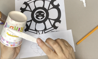 A pair of hands applying glue to a paper cup with a plastic knife. A flower drawing and design on the table.