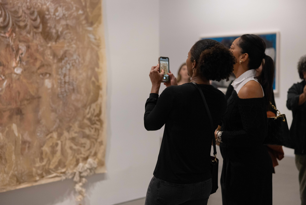 Two visitors look at a hanging brown painting and take a picture of it