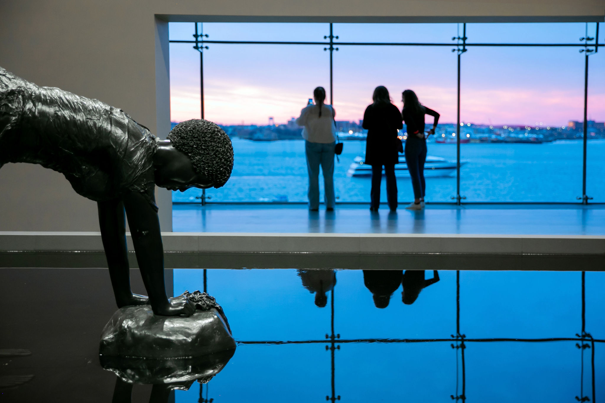 Bronze sculpture of woman bending down in pool of water with view of harbor at sunset in background