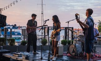 A band performs on an outdoor stage near a marina at sunset. Three musicians play guitar, bass, and drums, with microphones set up. Boats and string lights are visible in the background.