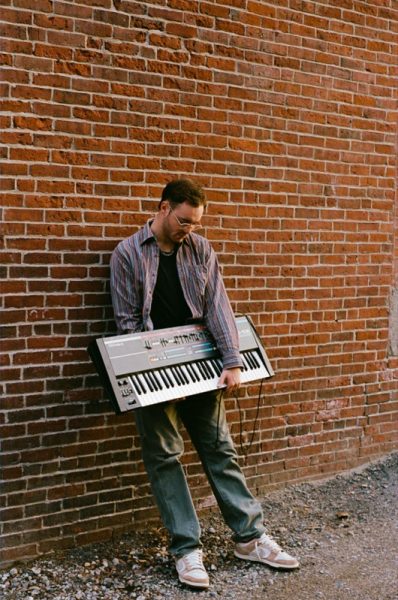 a man in jeans white shoes and a brown button up holds a keyboard and leans on a brick wall
