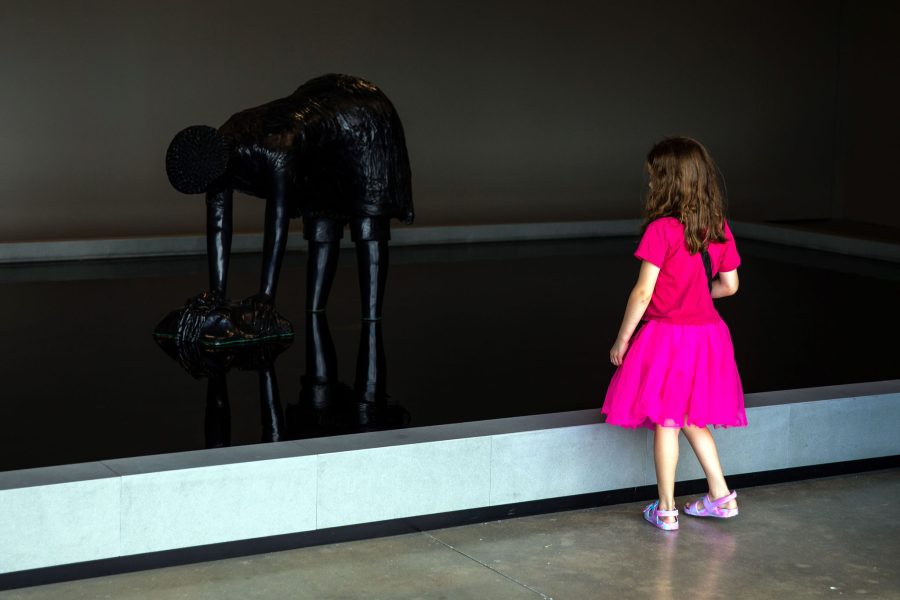 Child in bright pink dress looks at sculpture of a woman bending over in a reflective pool